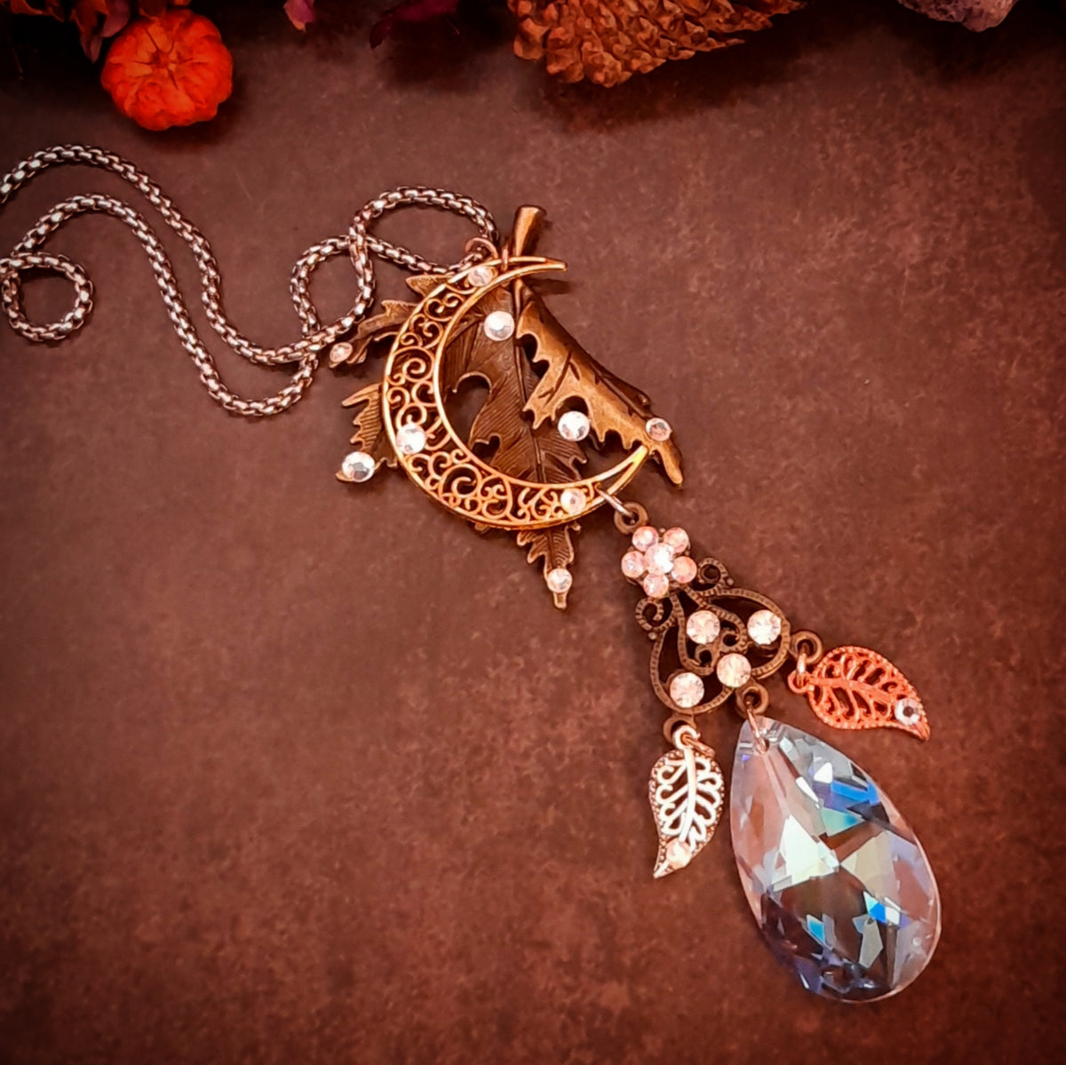 Handmade Victorian style and vintage style fantasy necklace by Rachel Flam Design, with a maple leaf, crescent moon, flower, leaf charms, and Swarovski® crystal teardrop on a brown background.