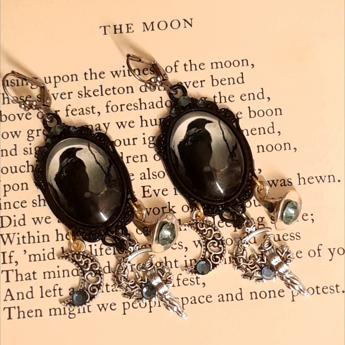 Witchy crow earrings photographed on an old book, featuring raven moon cabochons, smoky Swarovski crystals, crescent moon charms, witch figures, and celestial details, Victorian style and vintage style fantasy jewelry by Rachel Flam Design