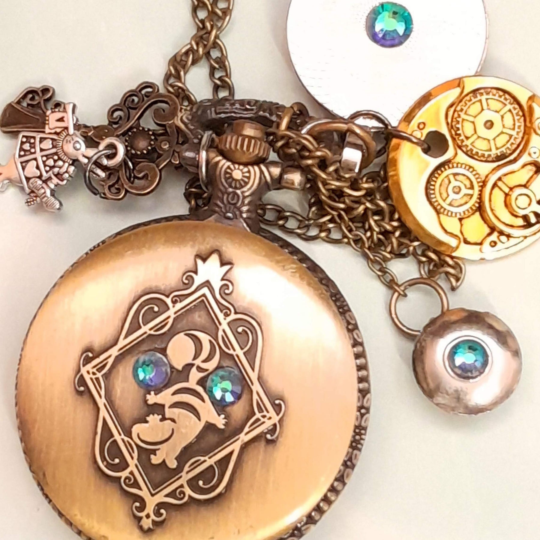 Alice in Wonderland pocket watch necklace back view showing Cheshire Cat engraving with steampunk gear and crystal disc charms antiqued brass Victorian style vintage style fantasy handmade swarovski crystals