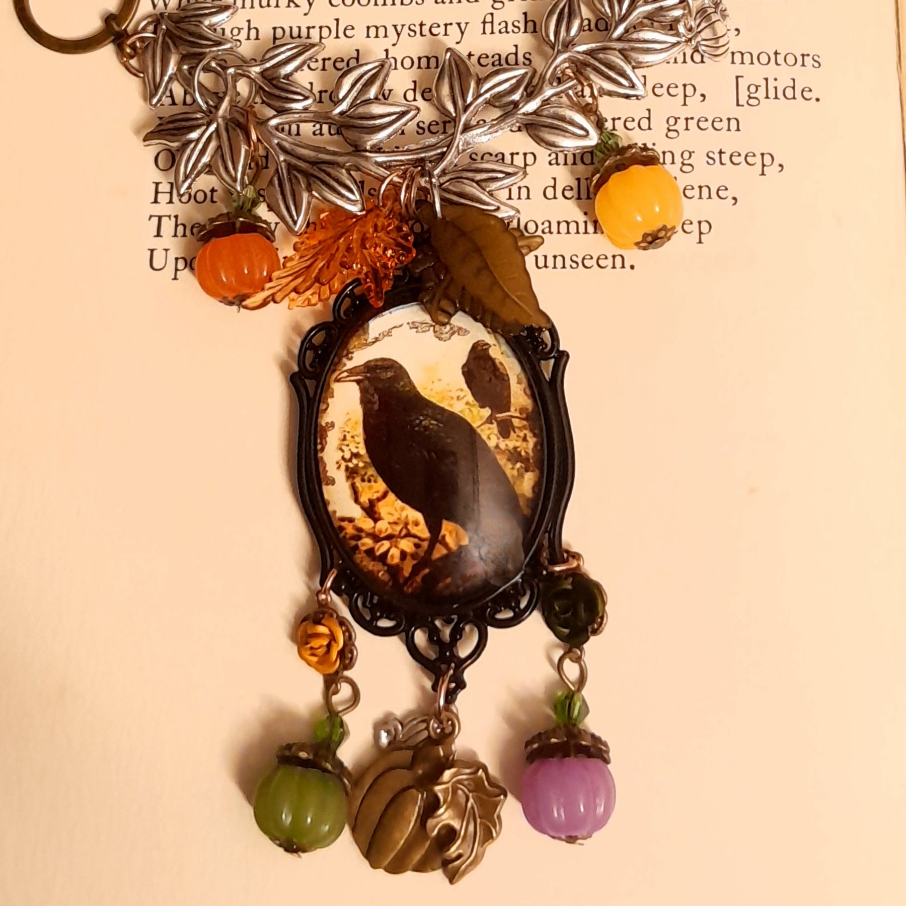 Black crow Halloween necklace photographed on an old book, featuring leafy branches, autumn leaves, pumpkin drops, raven artwork, and layered woodland details, Victorian style and vintage style fantasy jewelry by Rachel Flam Design