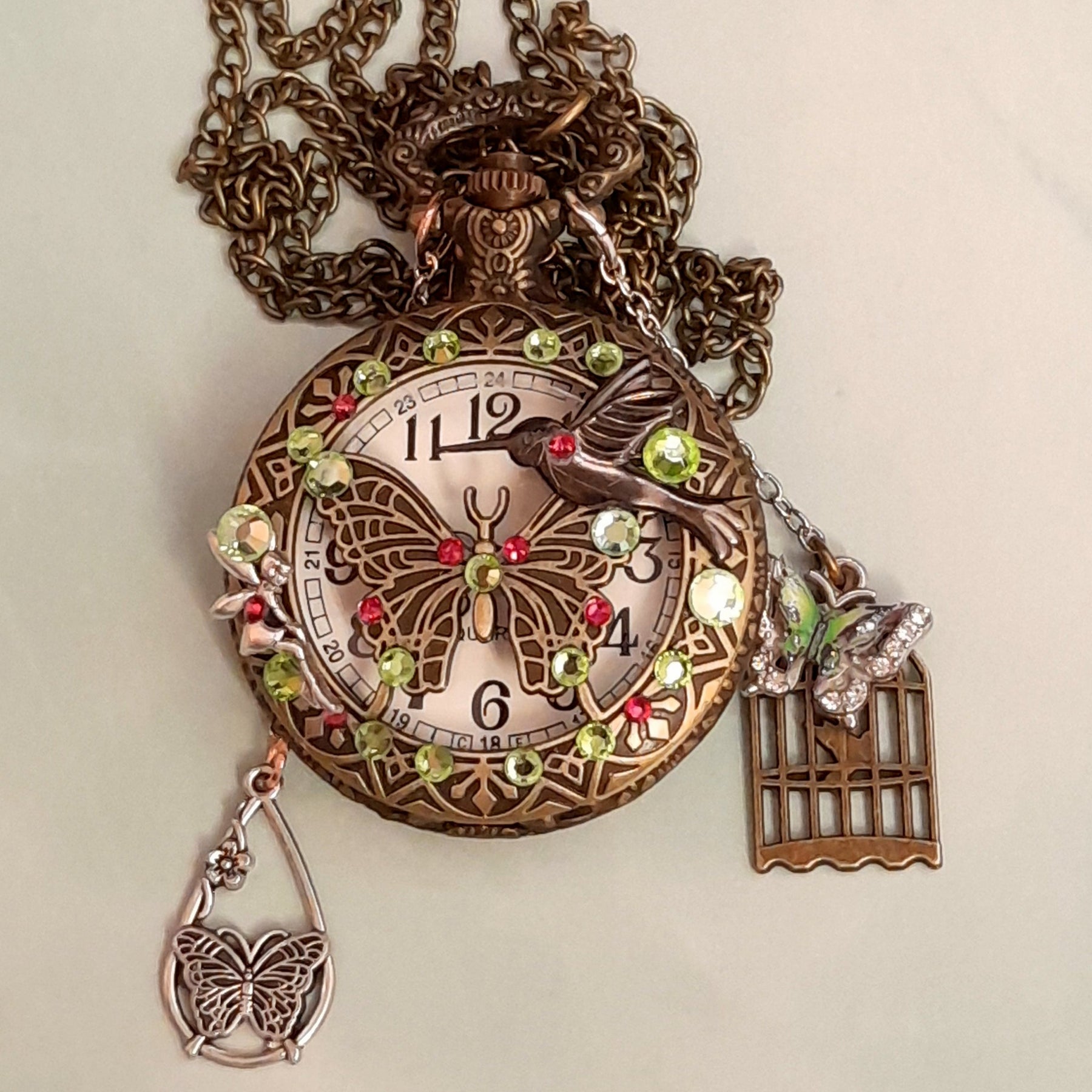 Handmade magical fantasy butterfly hummingbird pocketwatch necklace flat lay front view with peridot green and pink Swarovski crystals, butterfly birdcage charms, antiqued brass chain, Victorian style vintage style