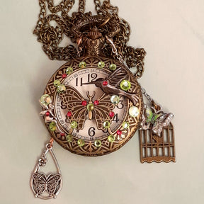 Handmade magical fantasy butterfly hummingbird pocketwatch necklace flat lay front view with peridot green and pink Swarovski crystals, butterfly birdcage charms, antiqued brass chain, Victorian style vintage style