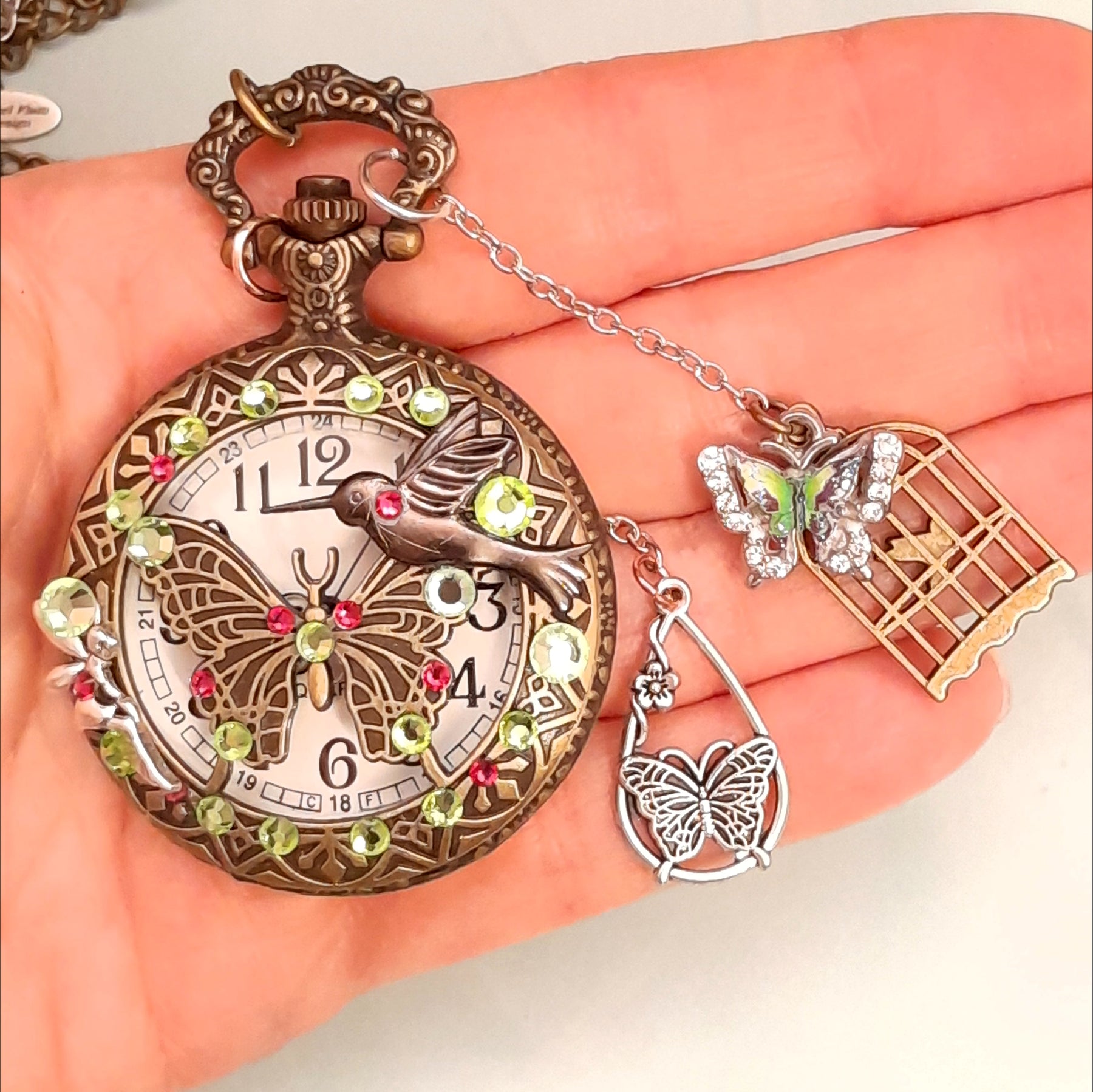 Handmade magical fantasy butterfly hummingbird pocketwatch necklace held in hand showing front with peridot green and pink Swarovski crystals, butterfly birdcage charms, antiqued brass, Victorian style vintage style