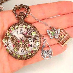 Handmade magical fantasy butterfly hummingbird pocketwatch necklace held in hand showing front with peridot green and pink Swarovski crystals, butterfly birdcage charms, antiqued brass, Victorian style vintage style