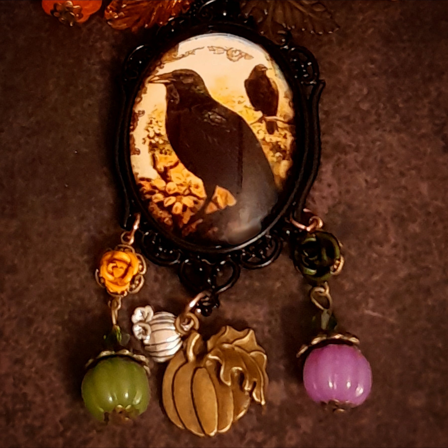 Close-up of handmade crow and pumpkin necklace showing the raven cabochon, autumn foliage, roses, pumpkin charms, and colorful Swarovski crystal accents, Victorian style and vintage style magical fantasy jewelry by Rachel Flam Design