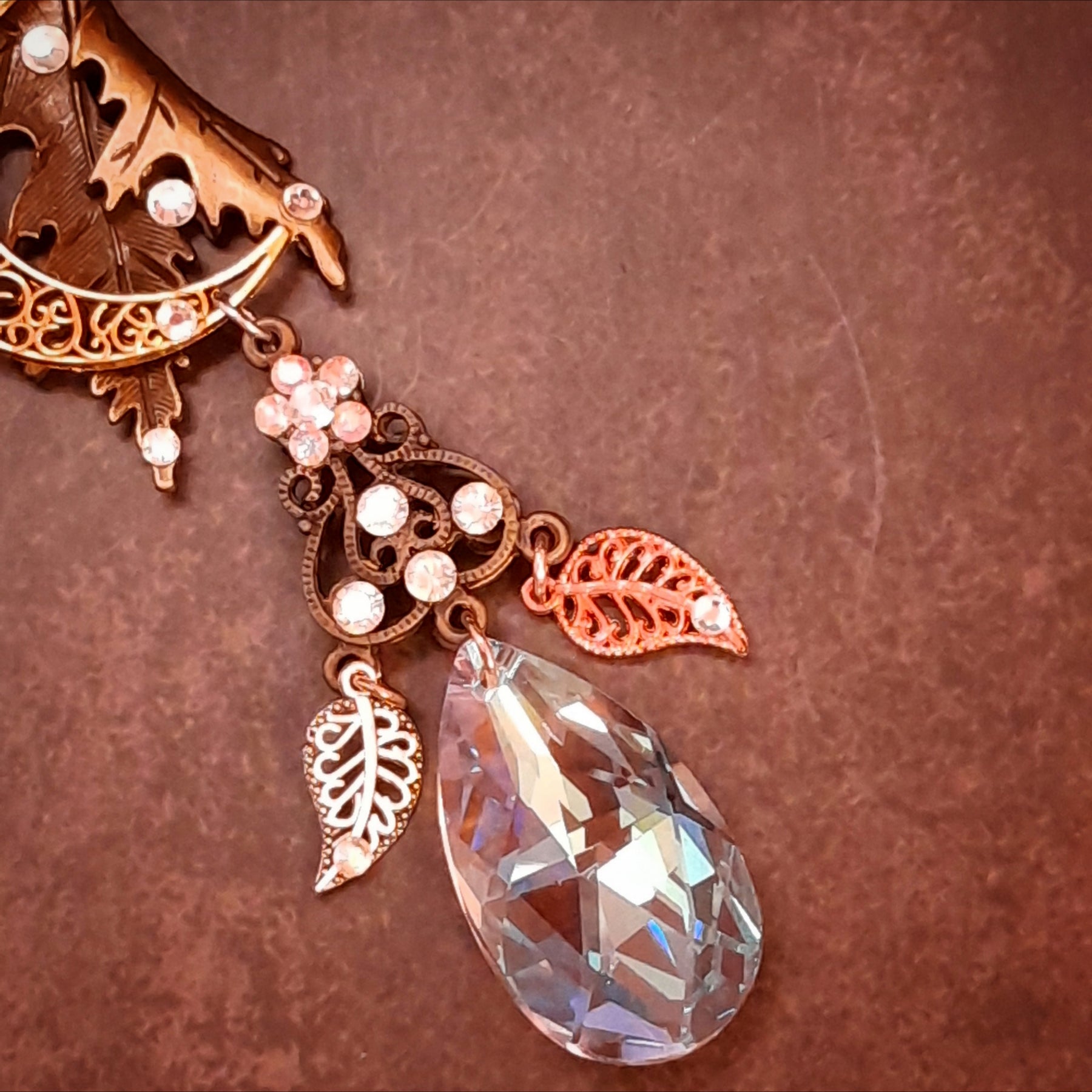 Close view of a faceted Swarovski® crystal teardrop between two openwork leaf charms on a Rachel Flam Design Victorian style and vintage style fantasy necklace, with a crystal flower above.
