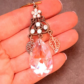 Close view of a Rachel Flam Design Victorian style and vintage style fantasy necklace held in a hand, showing a Swarovski® crystal teardrop, crystal flower, ornate connector, and two openwork leaf charms.