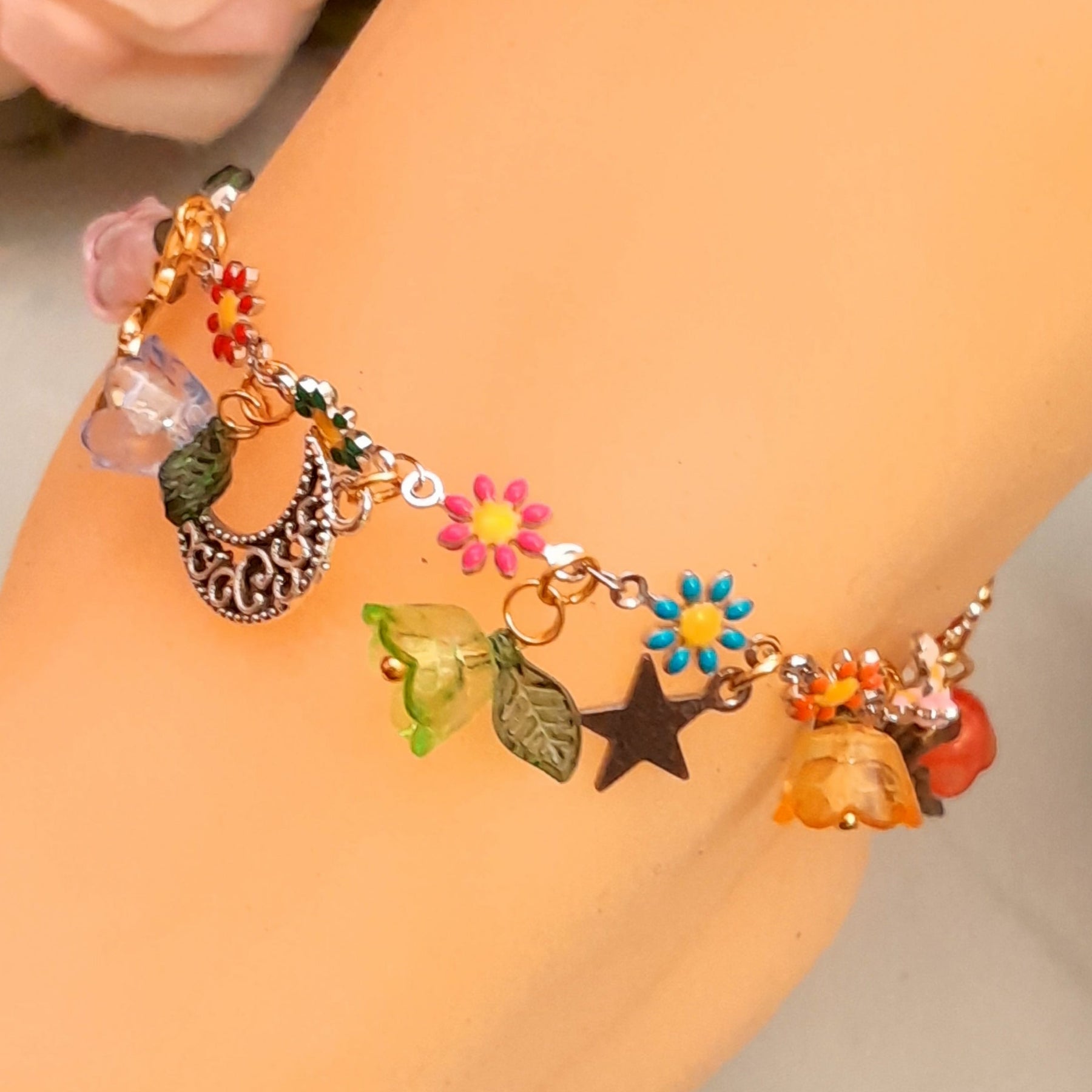 Handmade celestial fairy charm anklet worn on ankle featuring fairy figure, ornate crescent moon, star charm, multicolor enamel daisy flowers, and colorful flower beads with leaf accents, Victorian style vintage style