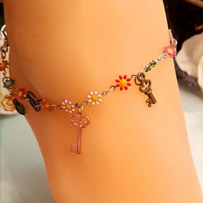 Closeup of handmade rainbow steampunk anklet showing gear cog charms, vintage skeleton keys, multicolor enamel daisy flowers, green crystal drops, and leaf charms, Victorian style vintage style