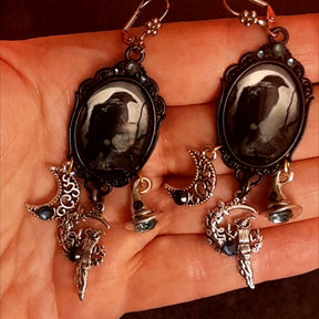 Handmade crow dangle earrings shown in hand with pale moon raven cabochons, smoky crystals, crescent moon charms, and detailed witch figures, Victorian style and vintage style fantasy jewelry by Rachel Flam Design