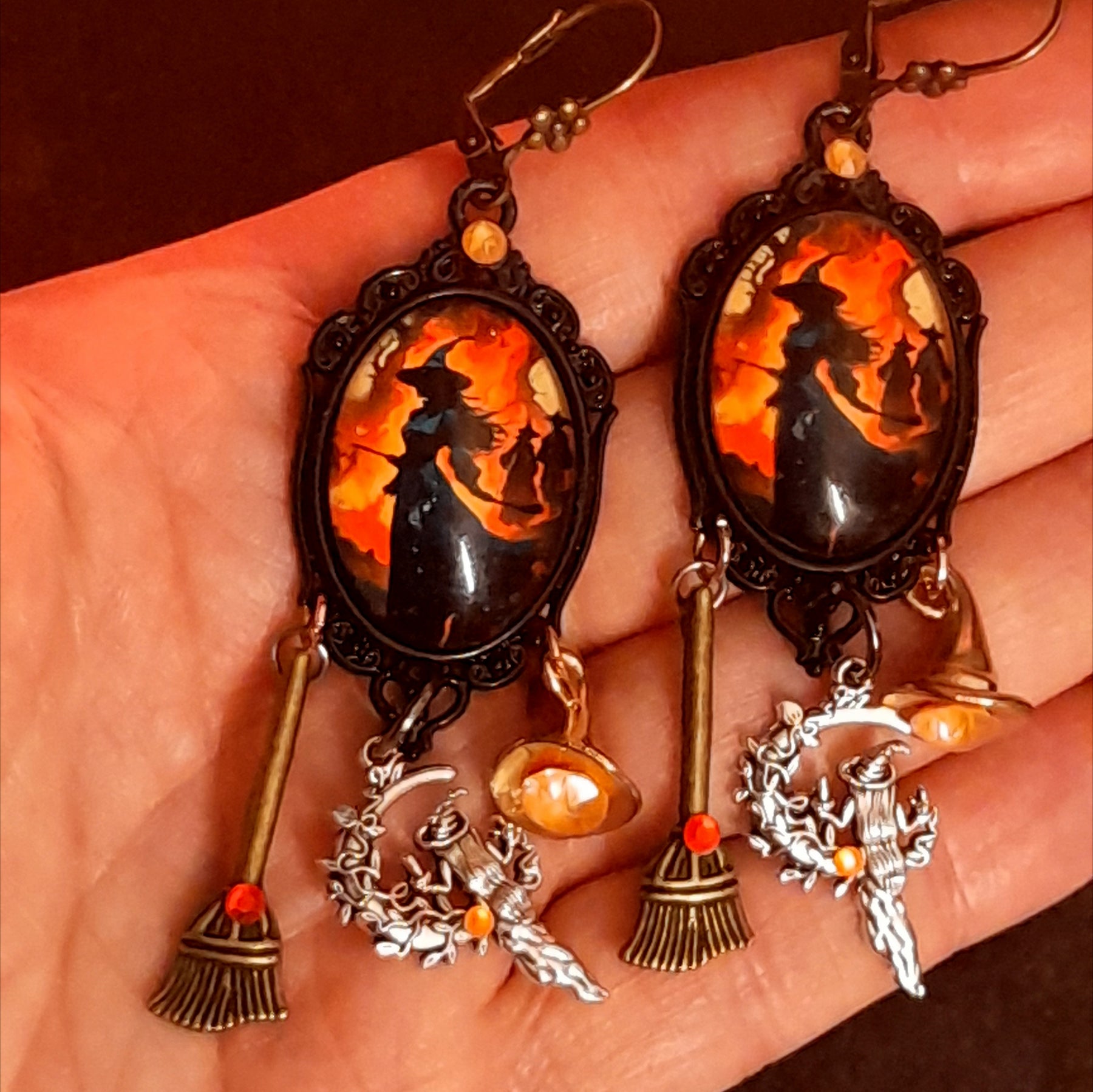 Handmade witchy earrings shown in hand with glowing orange witch artwork, red and orange Swarovski crystals, broom charms, witch hat details, and hidden fairy accents, Victorian style and vintage style magical fantasy jewelry by Rachel Flam Design