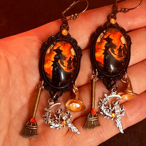 Handmade witchy earrings shown in hand with glowing orange witch artwork, red and orange Swarovski crystals, broom charms, witch hat details, and hidden fairy accents, Victorian style and vintage style magical fantasy jewelry by Rachel Flam Design