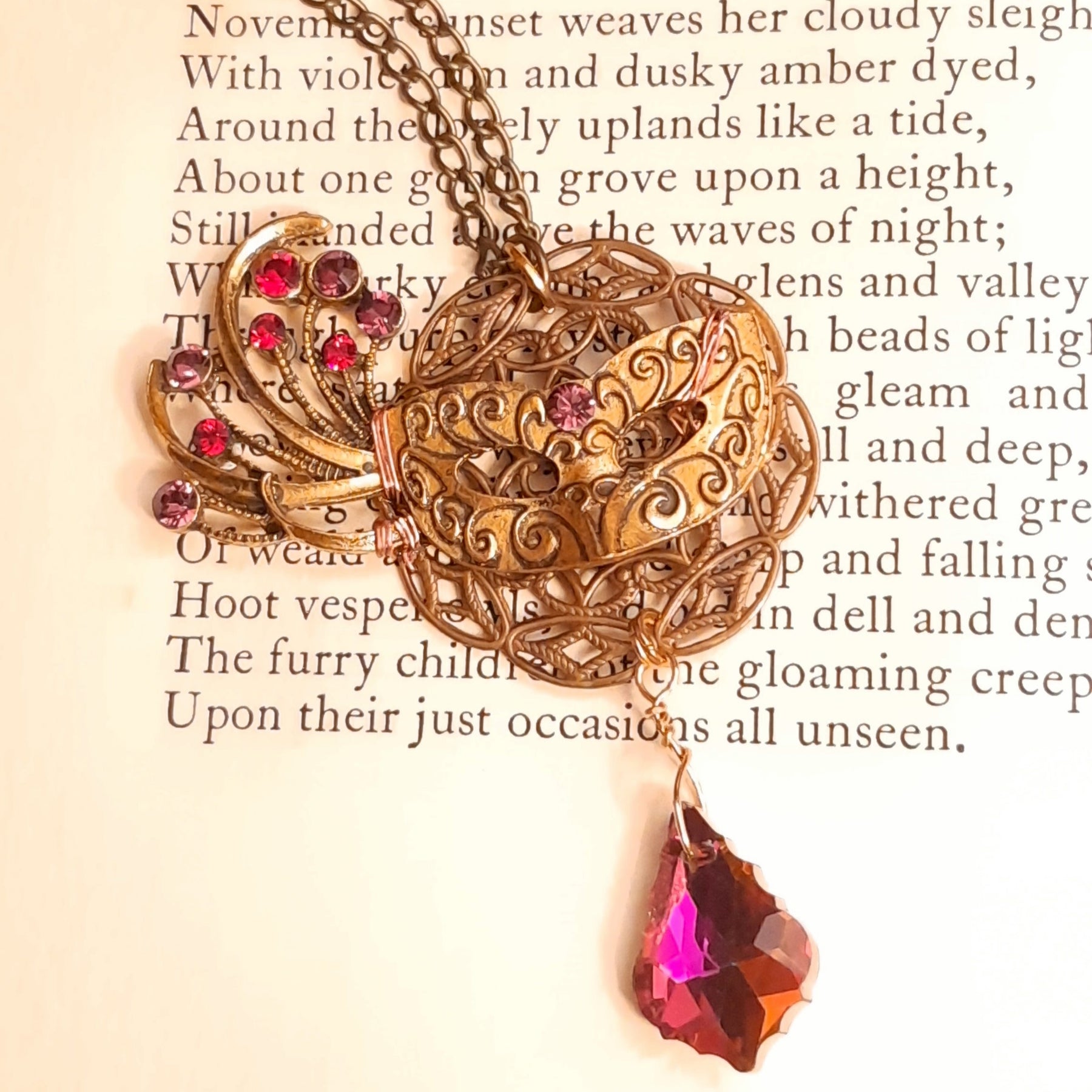 Victorian style vintage style masquerade mask necklace pendant resting on an antique poetry book page beside its red and pink Swarovski crystal feather fan