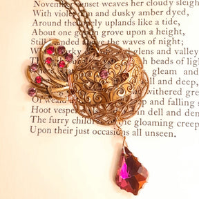 Victorian style vintage style masquerade mask necklace pendant resting on an antique poetry book page beside its red and pink Swarovski crystal feather fan
