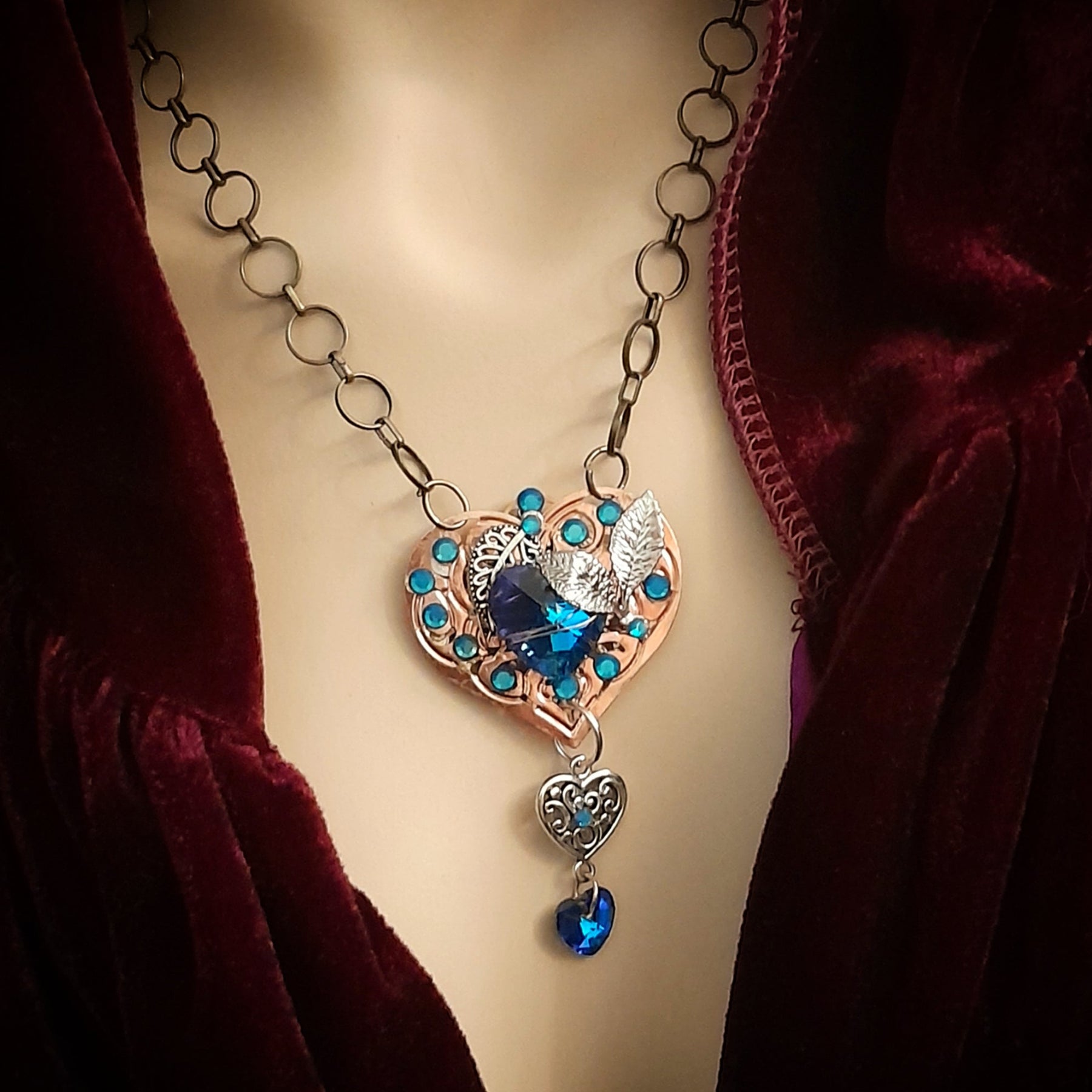 A necklace featuring a laser-engraved copper heart with blue crystals, silver leaves, and a large blue crystal at the center, displayed on a mannequin with a red velvet garment.