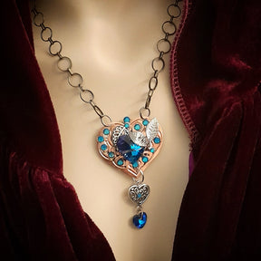 A necklace featuring a laser-engraved copper heart with blue crystals, silver leaves, and a large blue crystal at the center, displayed on a mannequin with a red velvet garment.