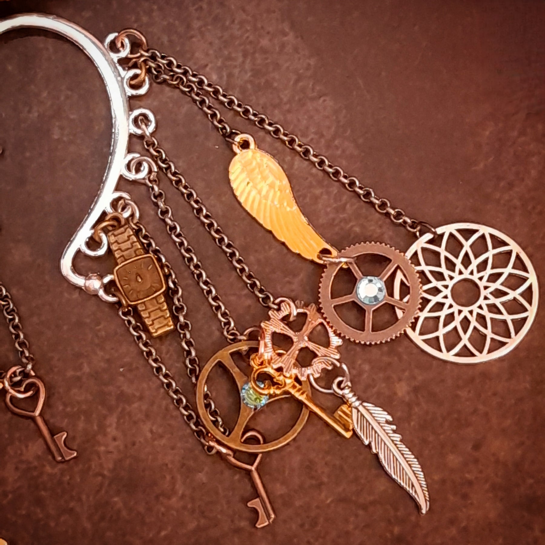 Detailed view of a Rachel Flam Design steampunk ear wrap and ear cuff embellished with light blue Swarovski® crystals, featuring a wing, feather, gears, keys, and an openwork circle in Victorian style and vintage style.