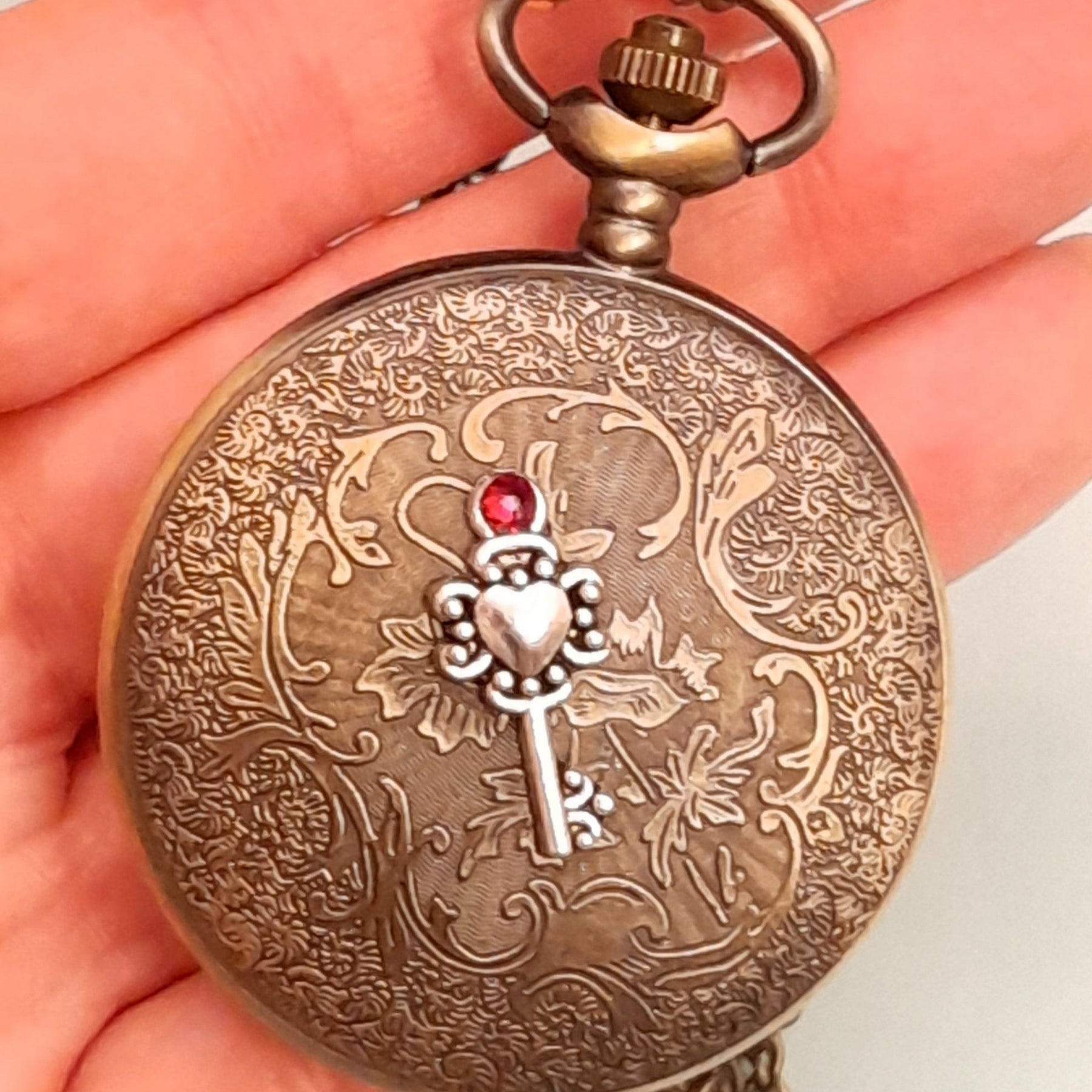 Steampunk pocket watch necklace held in hand closed f showing engraved floral design with skeleton key charm and red Swarovski crystal antiqued brass Victorian style vintage style fantasy handmadeback