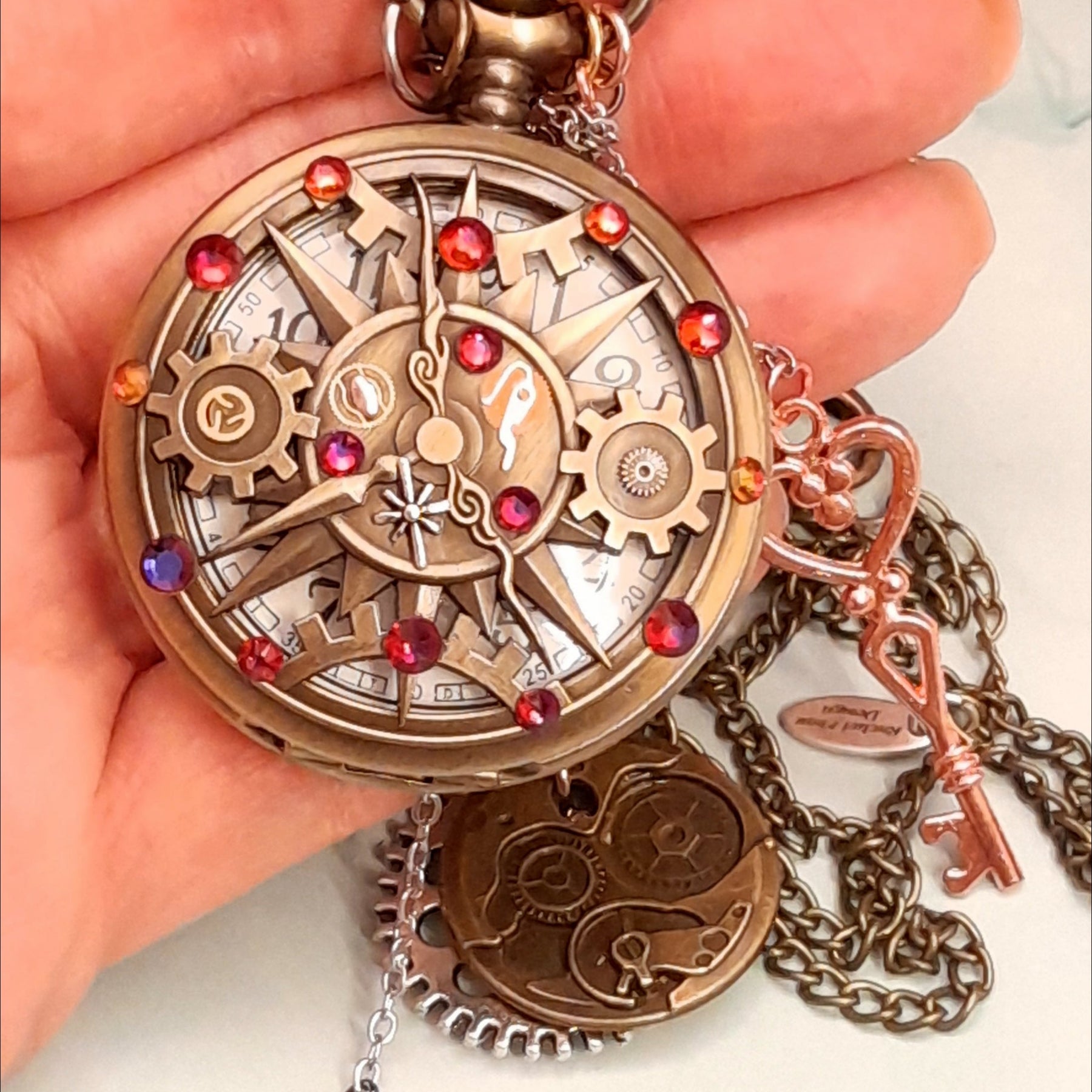 Steampunk pocket watch necklace held in hand closed front showing engraved floral design with skeleton key charm and red Swarovski crystal antiqued brass Victorian style vintage style fantasy handmade