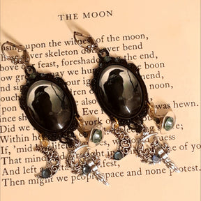 Witchy crow earrings photographed on an old book, featuring raven moon cabochons, smoky Swarovski crystals, crescent moon charms, witch figures, and celestial details, Victorian style and vintage style fantasy jewelry by Rachel Flam Design