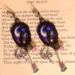 Witchy dangle earrings photographed on an old book, showing silhouetted witches against glowing crescent moons, purple Swarovski crystals, witch hat and broom details, Victorian style and vintage style fantasy jewelry by Rachel Flam Design