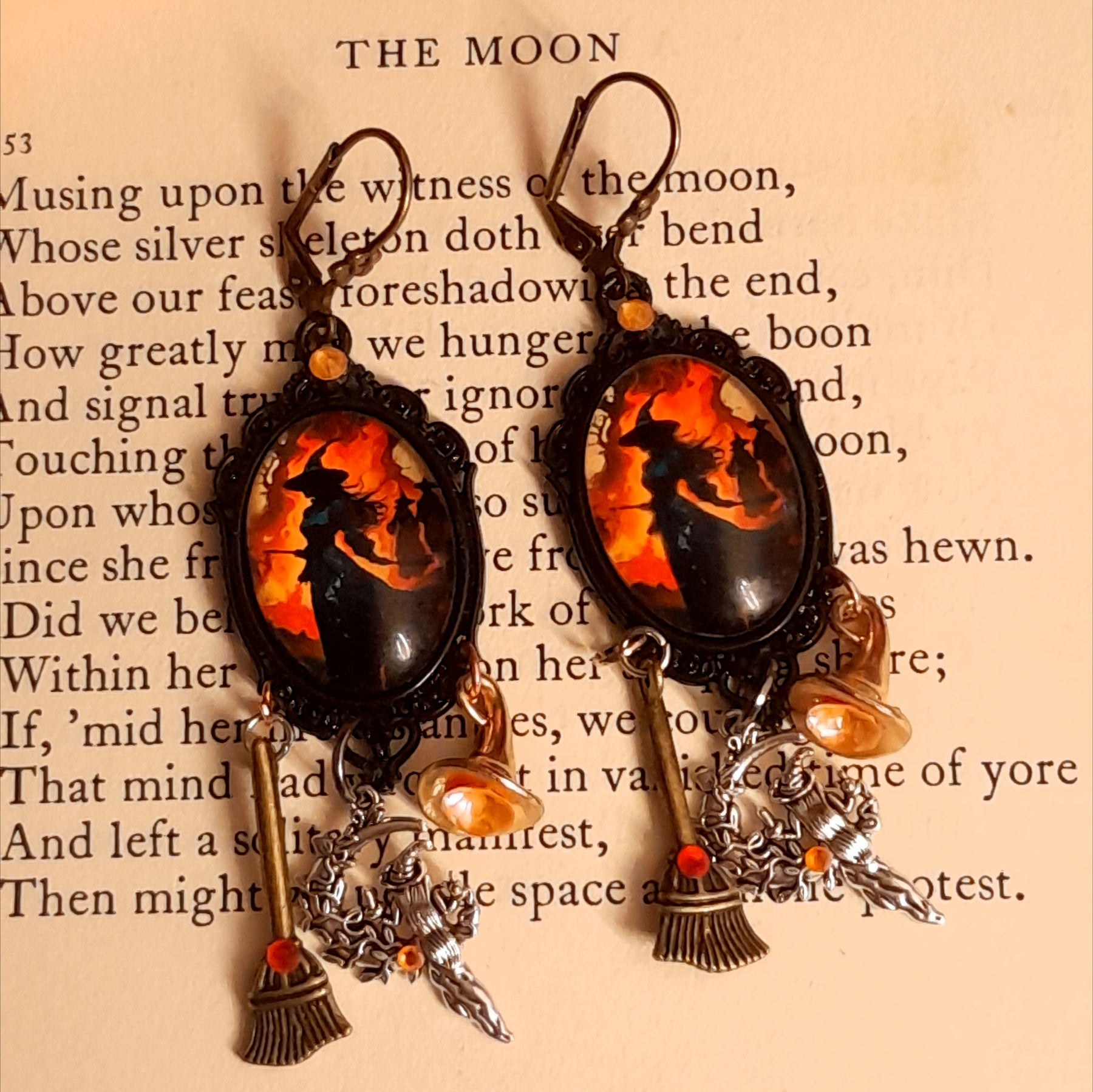 Witchy orange and red crystal earrings photographed on an old book, featuring silhouetted witch scenes, long broom dangles, crystal-filled witch hats, and celestial details, Victorian style and vintage style fantasy jewelry by Rachel Flam Design