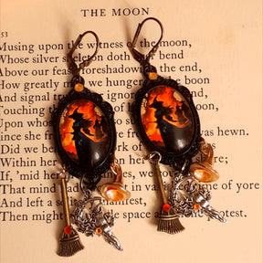 Witchy orange and red crystal earrings photographed on an old book, featuring silhouetted witch scenes, long broom dangles, crystal-filled witch hats, and celestial details, Victorian style and vintage style fantasy jewelry by Rachel Flam Design