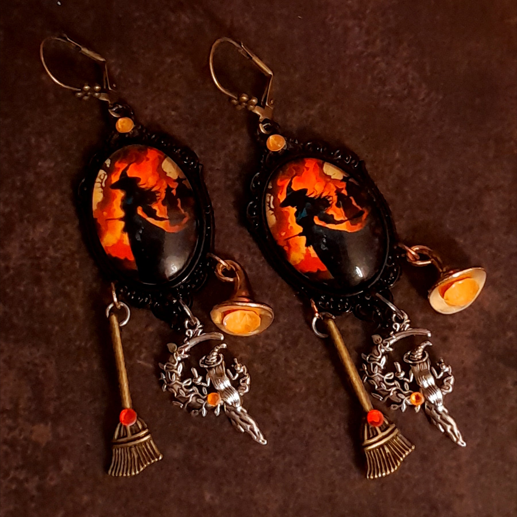 Handmade witchy earrings with glowing orange Swarovski crystals, firelit witch artwork, broom dangles, witch hat details, and red crystal accents, Victorian style and vintage style magical fantasy jewelry by Rachel Flam Design
