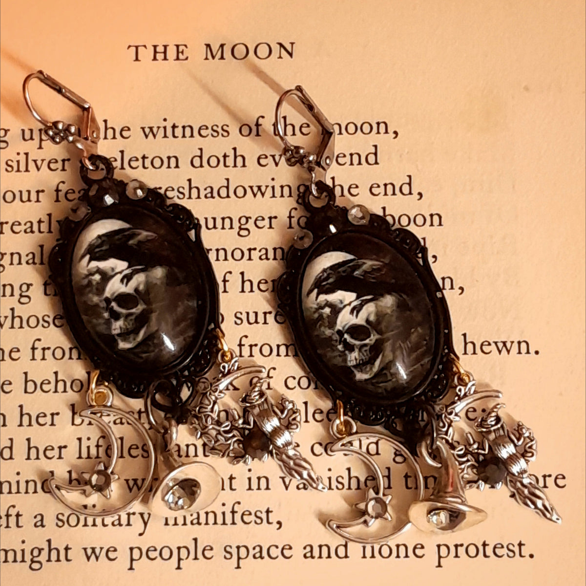 Witchy raven and skull earrings photographed on an old book, featuring smoky Swarovski crystals, crescent moons, witch hats, witch figures, and celestial charms, Victorian style and vintage style fantasy jewelry by Rachel Flam Design