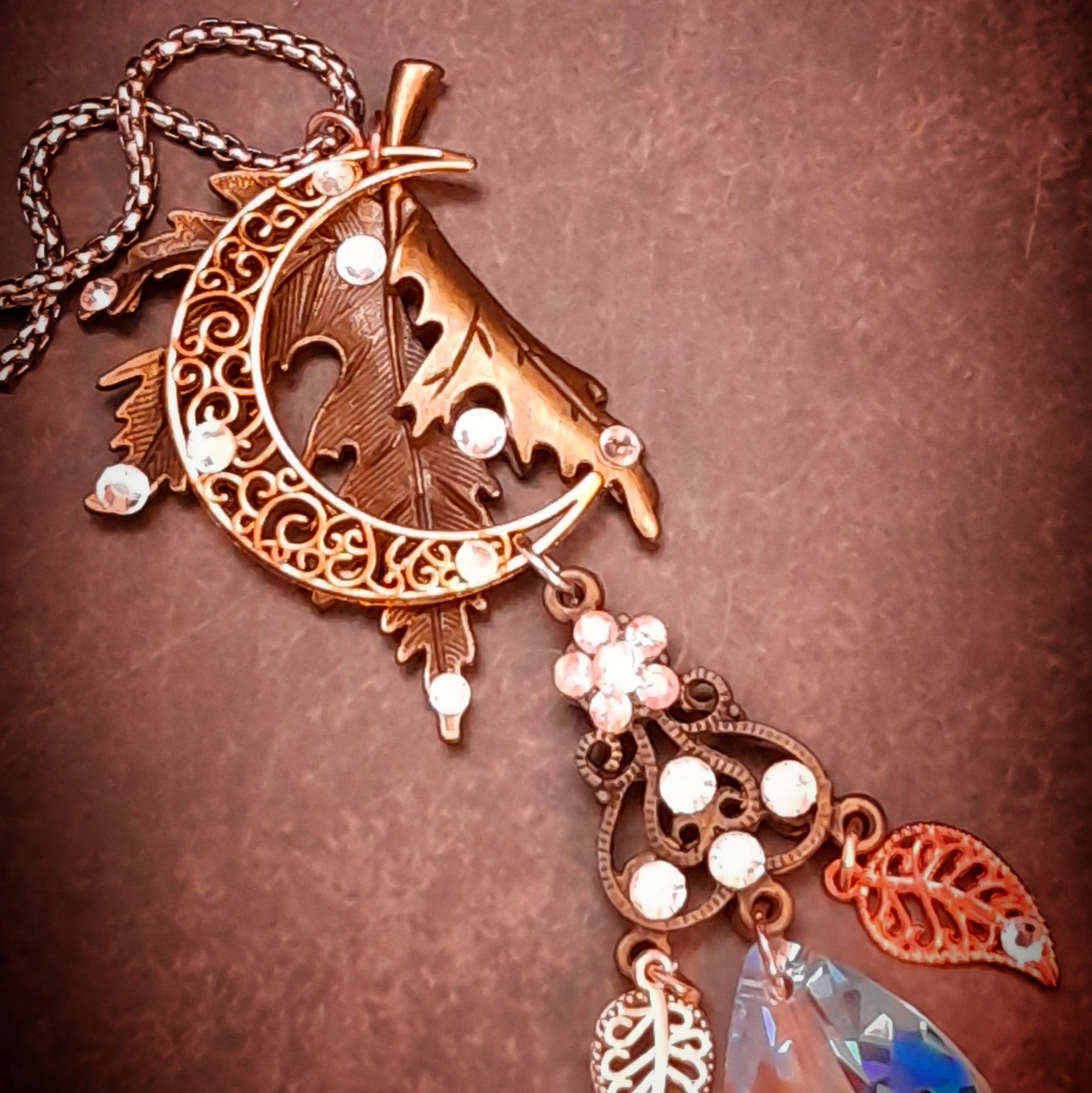 Rachel Flam Design Victorian style and vintage style woodland fantasy necklace on a brown background beneath a pinecone, featuring a maple leaf, crescent moon, and Swarovski® crystal accents.