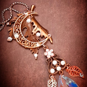 Rachel Flam Design Victorian style and vintage style woodland fantasy necklace on a brown background beneath a pinecone, featuring a maple leaf, crescent moon, and Swarovski® crystal accents.