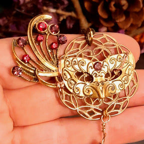 Victorian style vintage style masquerade mask necklace pendant held in hand showing the filigree face and red and pink Swarovski crystal feather fan detail